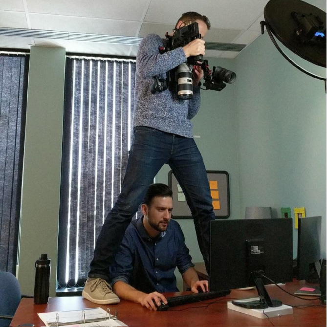 Dan Schuchman standing on an office desk while filming someone using a computer
